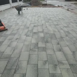 Ułożona kostka brukowa w odcieniach szarości na podjeździe, widoczny wózek budowlany, ubijarka, oraz przygotowany stos materiałów, w tle ulica i zabudowa jednorodzinna.