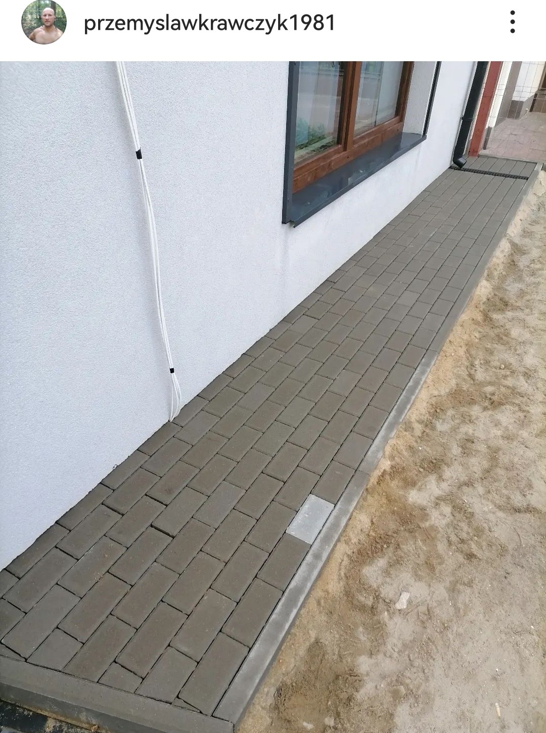 Ułożona kostka brukowa przy ścianie budynku z oknem, widoczny fragment rynny i kabli na elewacji, jedna kostka w innym kolorze.