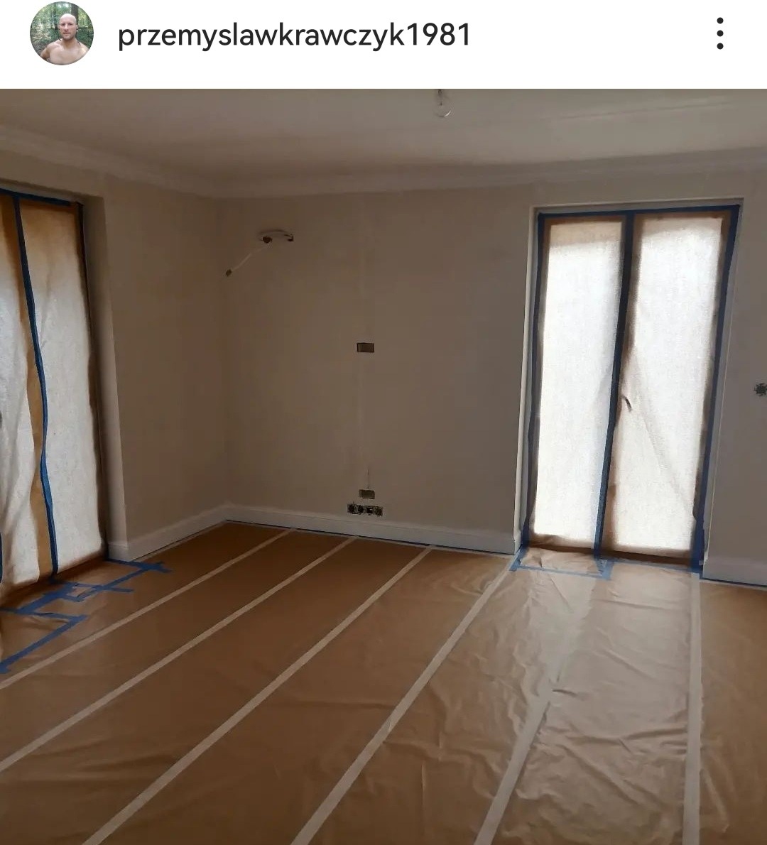 Pokój przygotowany do malowania: podłoga zabezpieczona folią malarską z taśmą, okna i drzwi osłonięte tkaniną, ściany w jasnym kolorze z widocznymi gniazdkami elektrycznymi.