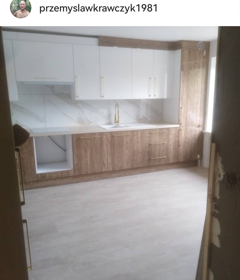 Nowoczesna kuchnia z białymi szafkami górnymi i drewnopodobnymi dolnymi, złota armatura, marmurowe płytki na ścianie, jasna podłoga, widok zza framugi drzwi.