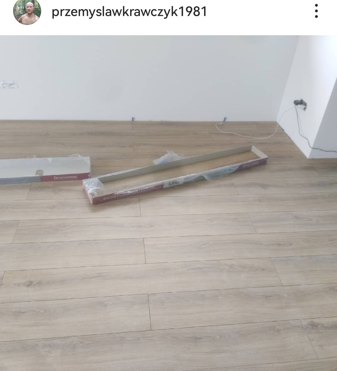 Jasne panele podłogowe z widocznymi opakowaniami producentów 'Fachowiec' i 'Swiss Floor Collection' przed montażem w rogu pomieszczenia z białymi ścianami.