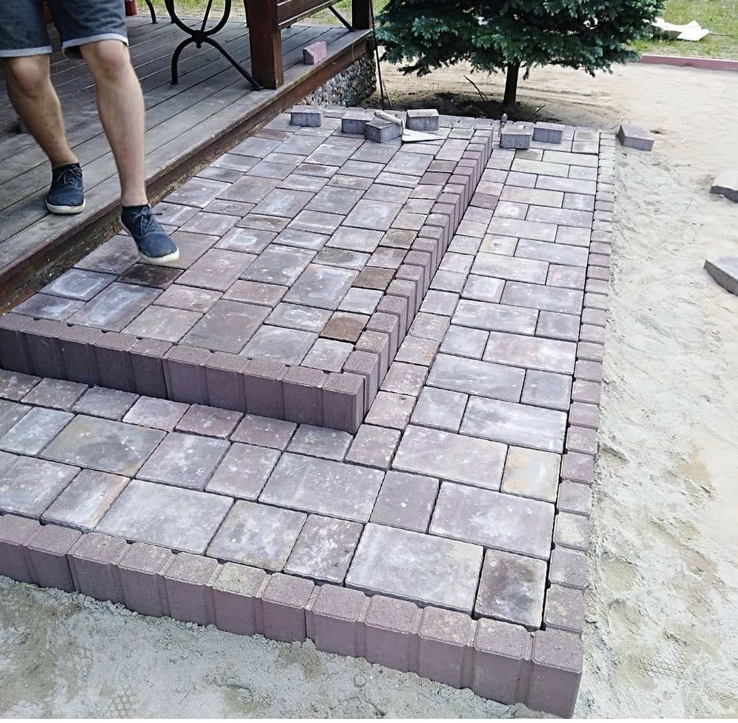 Ułożona kostka brukowa tworząca schody wejściowe na taras, widoczne fragmenty osoby w szortach i tenisówkach, piasek wokół nowej konstrukcji.