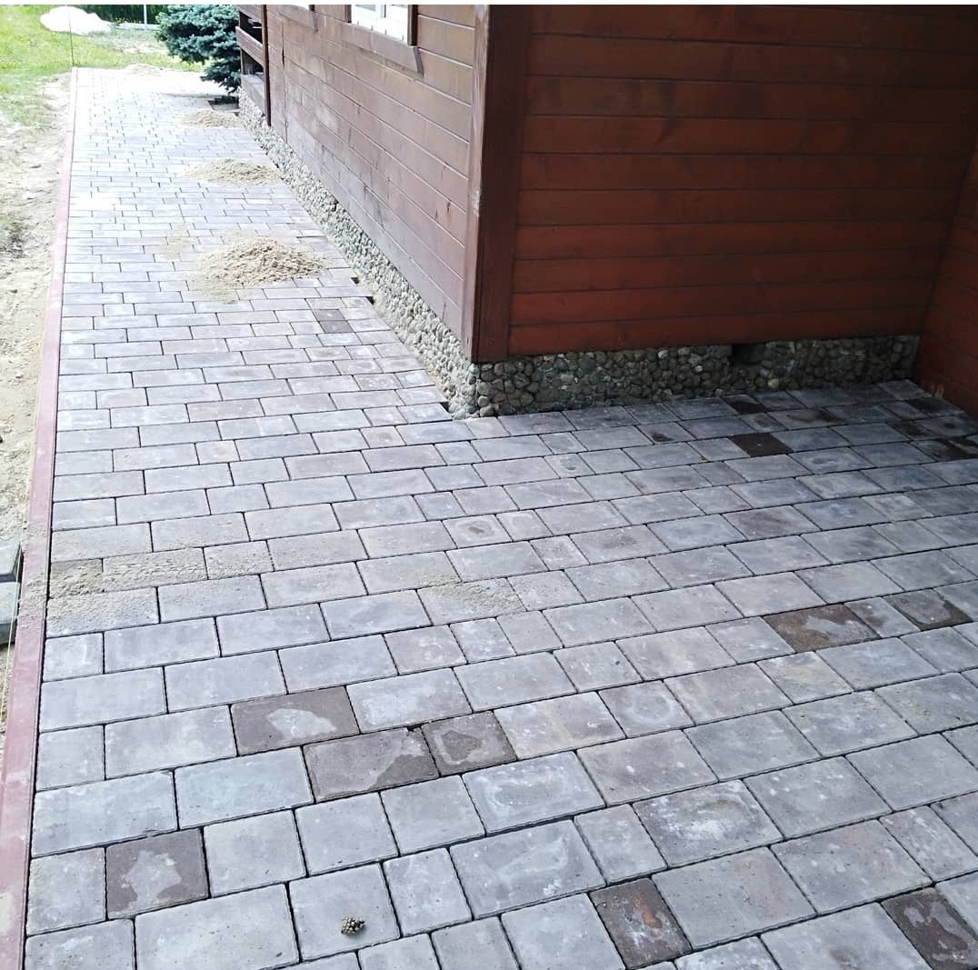 Ułożona kostka brukowa w odcieniach szarości i brązu tworząca ścieżkę przy drewnianym domu z podmurówką z kamieni, widoczne pozostałości piasku na kostce.