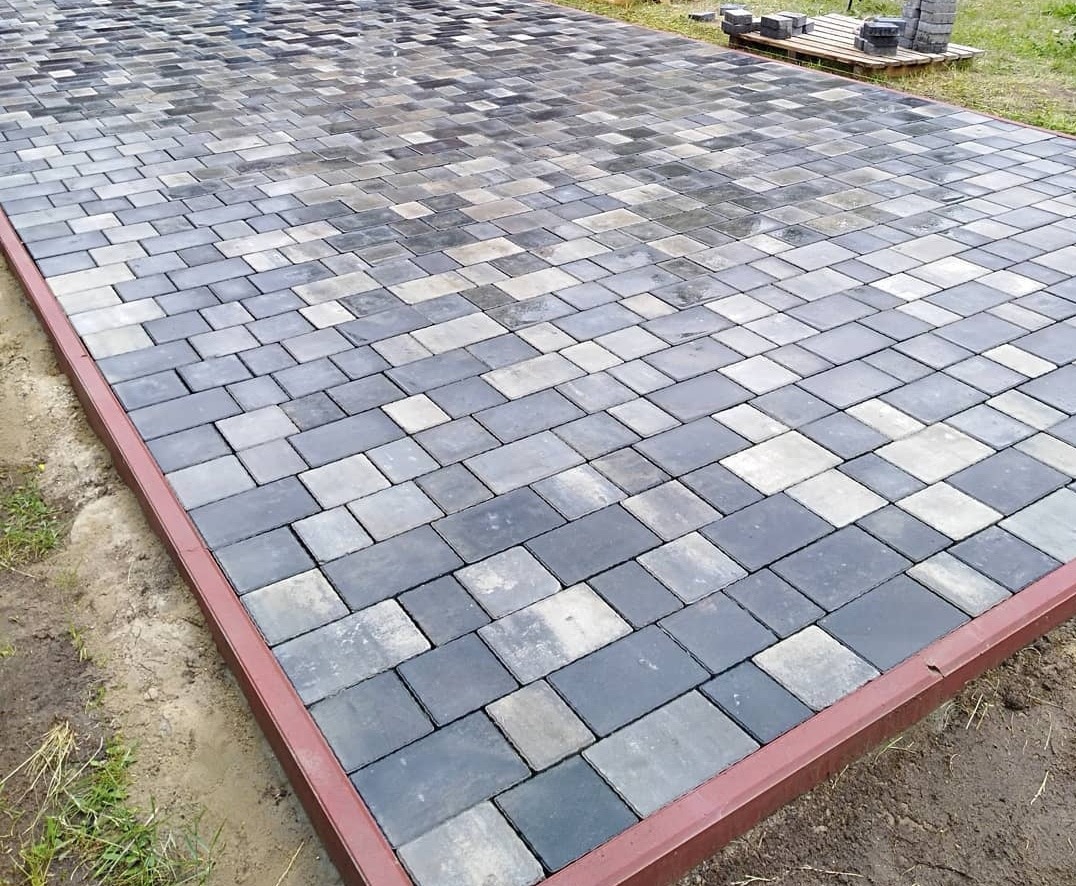 Ułożona kostka brukowa w odcieniach szarości i grafitu, obramowana czerwonobrązową metalową listwą. Częściowo mokra powierzchnia odbija światło, w tle paleta z elementami kostki.