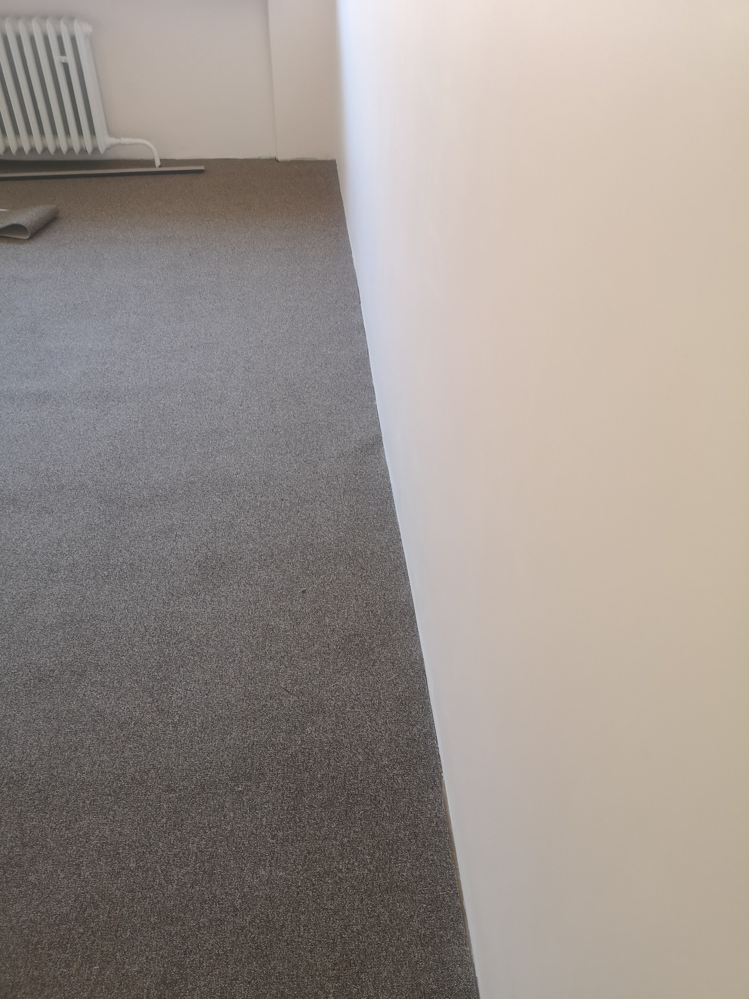 Ułożona szara wykładzina dywanowa w pomieszczeniu, widoczny fragment grzejnika i fragment zwiniętej wykładziny przy ścianie.