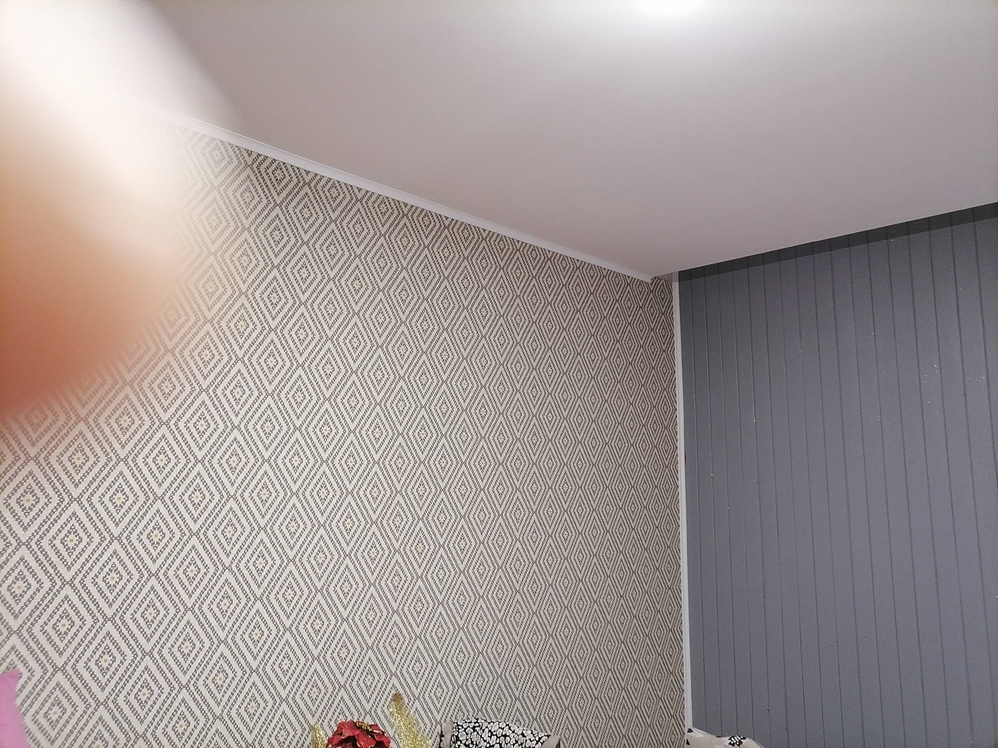 Wnętrze pokoju z tapetą w geometryczne wzory i szarą ścianą z paneli pionowych; widoczny fragment sufitu i dekoracje na podłodze.