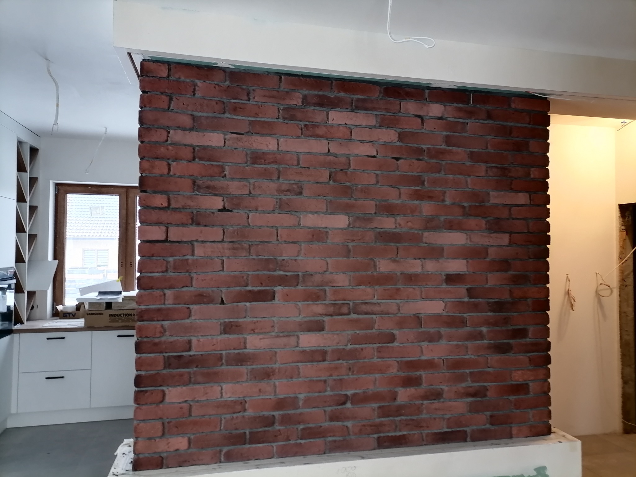 Ceglana ściana działowa w surowym stylu, oddzielająca kuchnię od innego pomieszczenia, widoczne niedokończone obróbki, wystające przewody elektryczne.