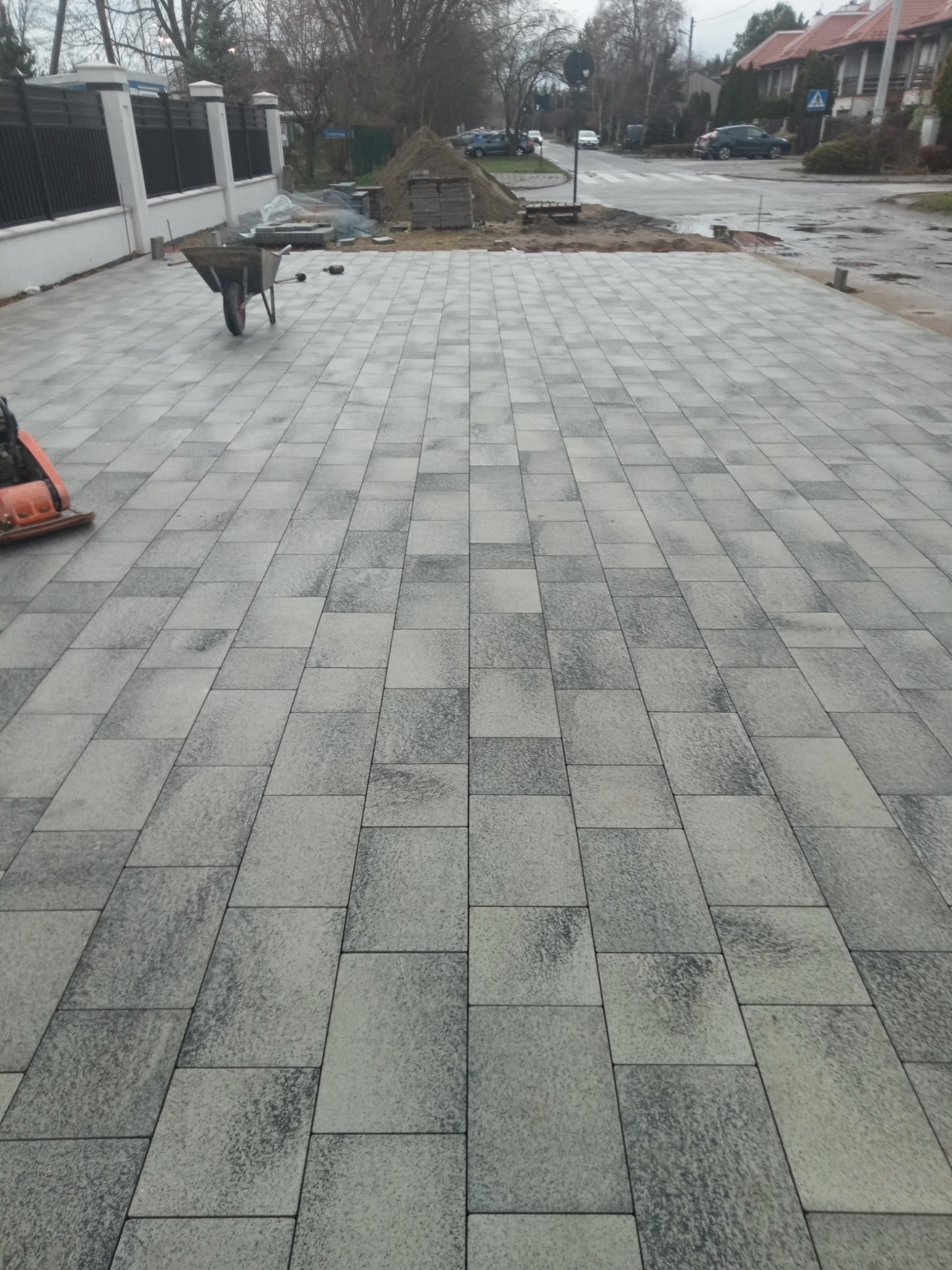 Ułożona kostka brukowa w odcieniach szarości na podjeździe, widoczny wózek budowlany, ubijarka, oraz przygotowany stos materiałów, w tle ulica i zabudowa jednorodzinna.