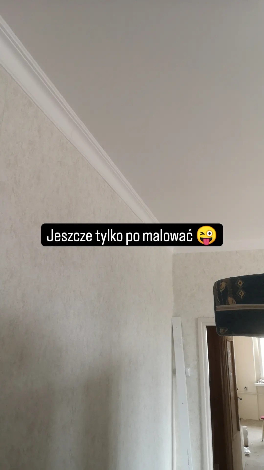 Ściana z fakturą i listwą dekoracyjną pod sufitem, przygotowana do malowania. Widoczny fragment futryny drzwi i element wyposażenia wnętrza w tle.