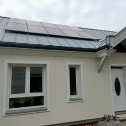 Dom jednorodzinny z zainstalowanymi panelami słonecznymi na metalowym dachu. Widoczne okna, drzwi wejściowe i elementy wykończeniowe budynku.