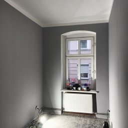 Maksym Sova Group Company - Pokój po malowaniu na szaro, widoczne ślady po remoncie na podłodze, grzejnik pod oknem, listwy przypodłogowe i karnisz pomalowane na biało.