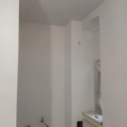 Wnętrze w trakcie remontu, świeżo pomalowane ściany na biało, widoczne instalacje hydrauliczne przed zabudową, okno z zabezpieczeniem folią.