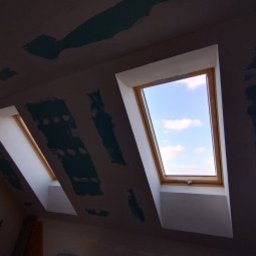 Maksym Sova Group Company - Montaż okien dachowych w skosie poddasza, widoczne łączenia płyt gipsowo-kartonowych przed szpachlowaniem, błękitne niebo przez okno.