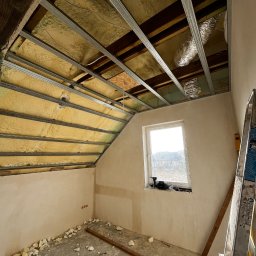 Maksym Sova Group Company - Montaż metalowej konstrukcji sufitu podwieszanego w skośnym pomieszczeniu z widoczną izolacją, fragmentem instalacji wentylacyjnej, oknem i narzędziami na parapecie oraz resztkami materiałów...