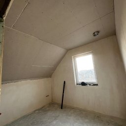Maksym Sova Group Company - Pokój na poddaszu w trakcie prac wykończeniowych, z zamontowaną płytą na skosach sufitu i ścianach, surowy beton na podłodze, poziomica oparta o ścianę obok okna.