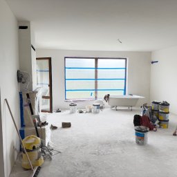 Maksym Sova Group Company - Wnętrze w trakcie malowania: zabezpieczone okno taśmą, drabina, puszki z farbą, narzędzia malarskie i odkurzacz. Ściany świeżo pomalowane na biało, widoczna wanna w tle.