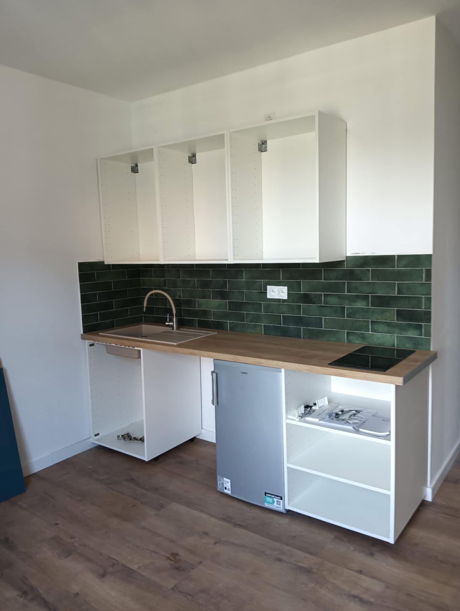 Jasna kuchnia z zieloną glazurą: białe szafki wiszące i dolne, drewniany blat, srebrny zlew i mała lodówka. Widoczne gniazdka elektryczne. Podłoga z paneli.