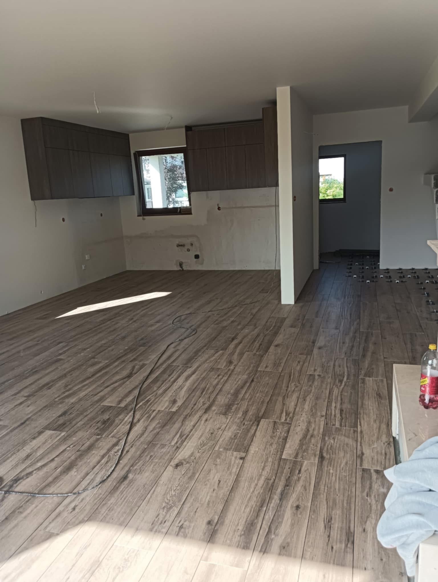 Układanie paneli podłogowych imitujących drewno w nowym wnętrzu. Widoczne płytki dystansowe, okno i fragmenty instalacji elektrycznej. Duże, jasne pomieszczenie w trakcie wykończenia.