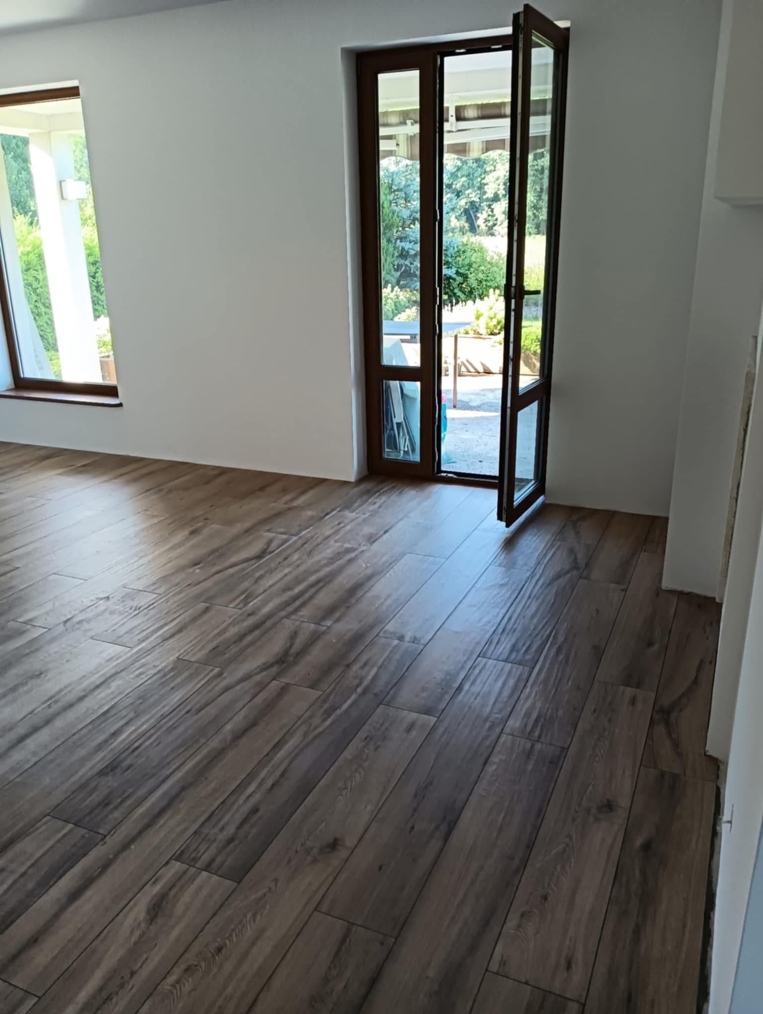 Ułożone panele podłogowe imitujące drewno w jasnym pokoju z oknem i otwartymi drzwiami balkonowymi, widok na zieleń na zewnątrz.