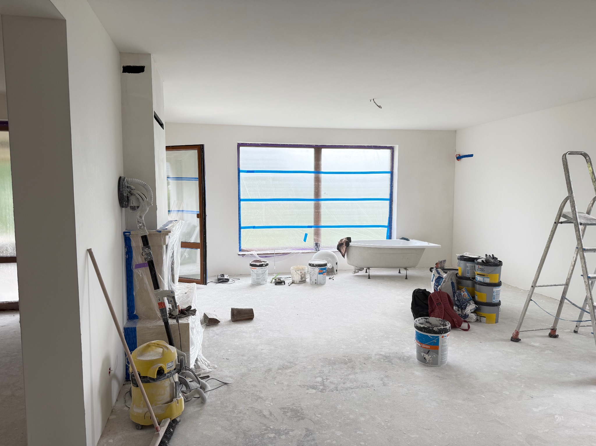Wnętrze w trakcie malowania: zabezpieczone okno taśmą, drabina, puszki z farbą, narzędzia malarskie i odkurzacz. Ściany świeżo pomalowane na biało, widoczna wanna w tle.