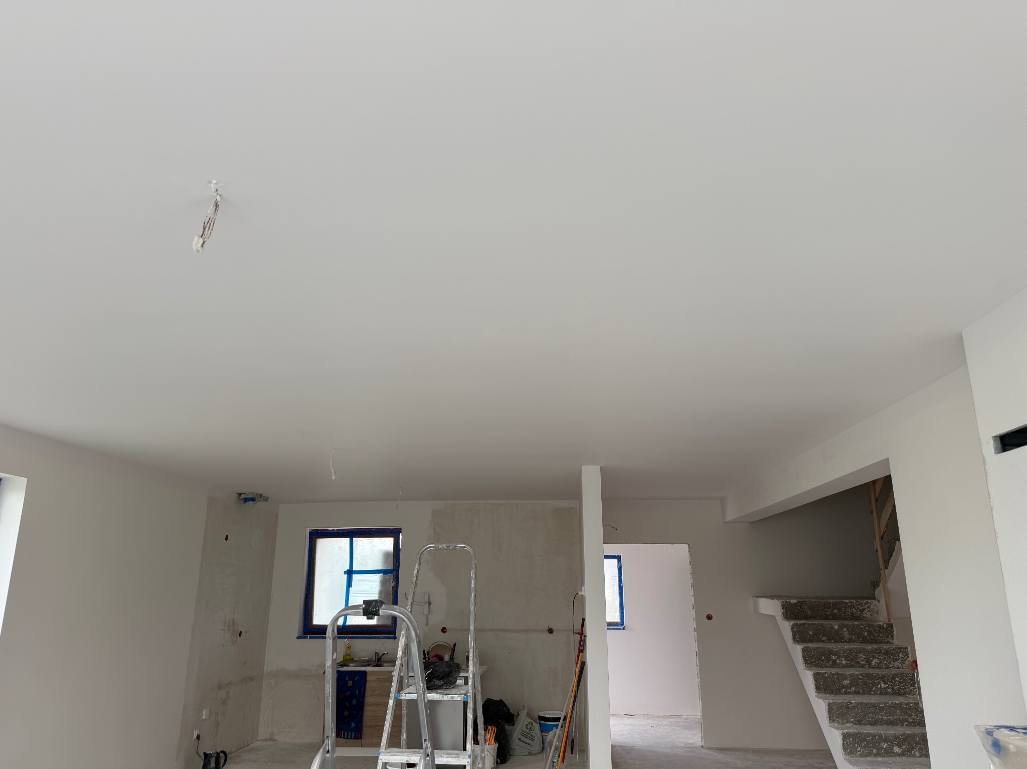 Wnętrze w trakcie remontu: białe ściany, okna z niebieską taśmą, drabina, schody i sufit z widocznymi śladami po instalacji. Widok na przestrzeń w trakcie odświeżania.