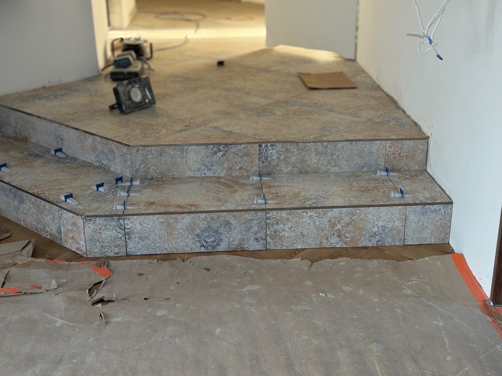 Wykończenie schodów płytkami z dekoracyjnym wzorem, widoczne niebieskie krzyżyki dystansowe. Podłoga zabezpieczona kartonem. W tle narzędzia i kable.