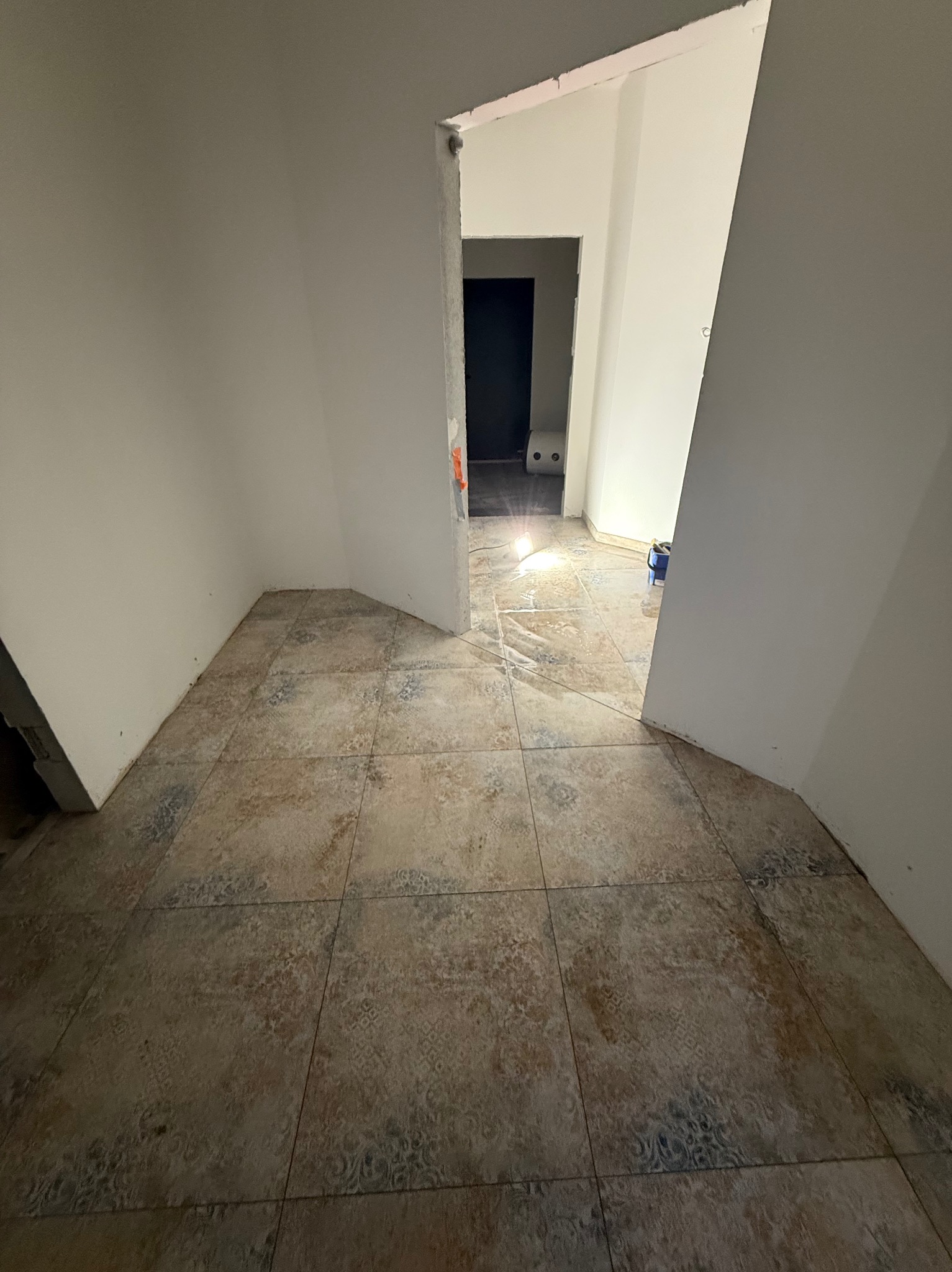 Wnętrze w trakcie remontu: świeżo pomalowane ściany na biało, wzorzysta podłoga z płytek. Widoczne wiadro i lampa halogenowa. Perspektywa z korytarza.