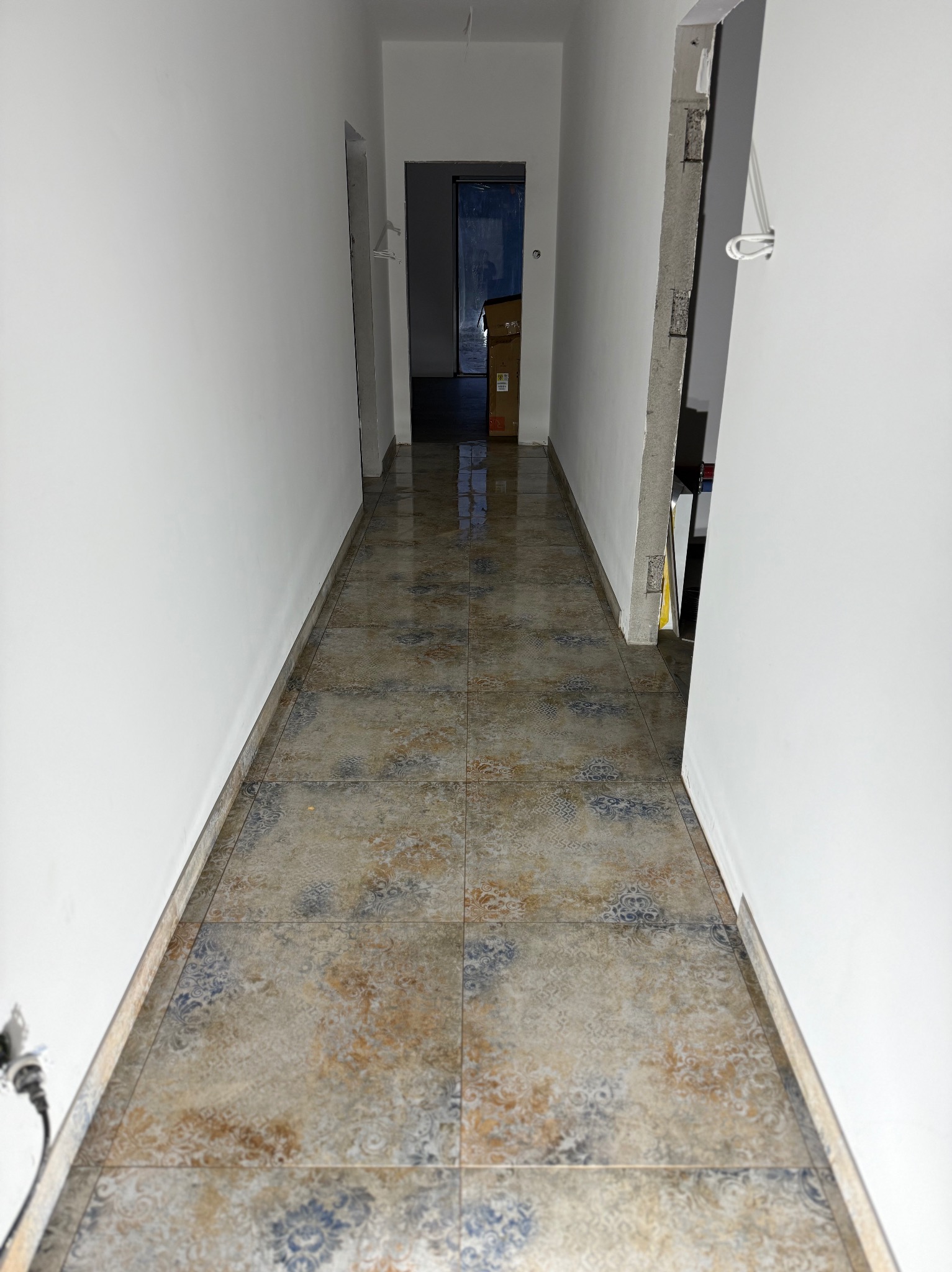 Korytarz po remoncie z nową, wzorzystą podłogą z płytek. Świeżo pomalowane, białe ściany. Widoczny fragment niezakończonego przejścia z wystającymi przewodami.