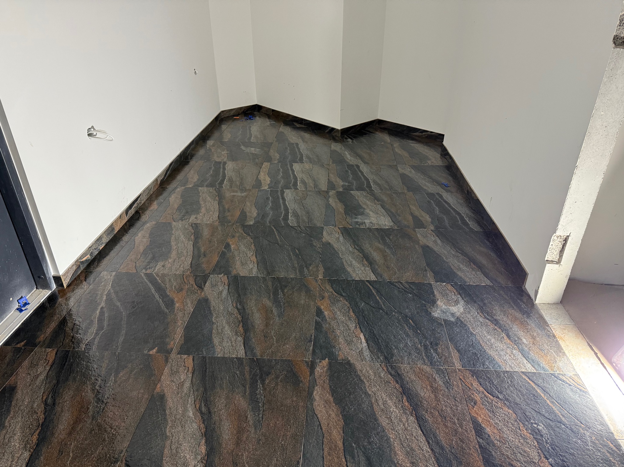 Ułożona, ciemna, błyszcząca podłoga z płytek imitujących kamień w rogu pomieszczenia. Widoczne białe ściany i fragment drzwi. Użyto niebieskich klinów poziomujących.