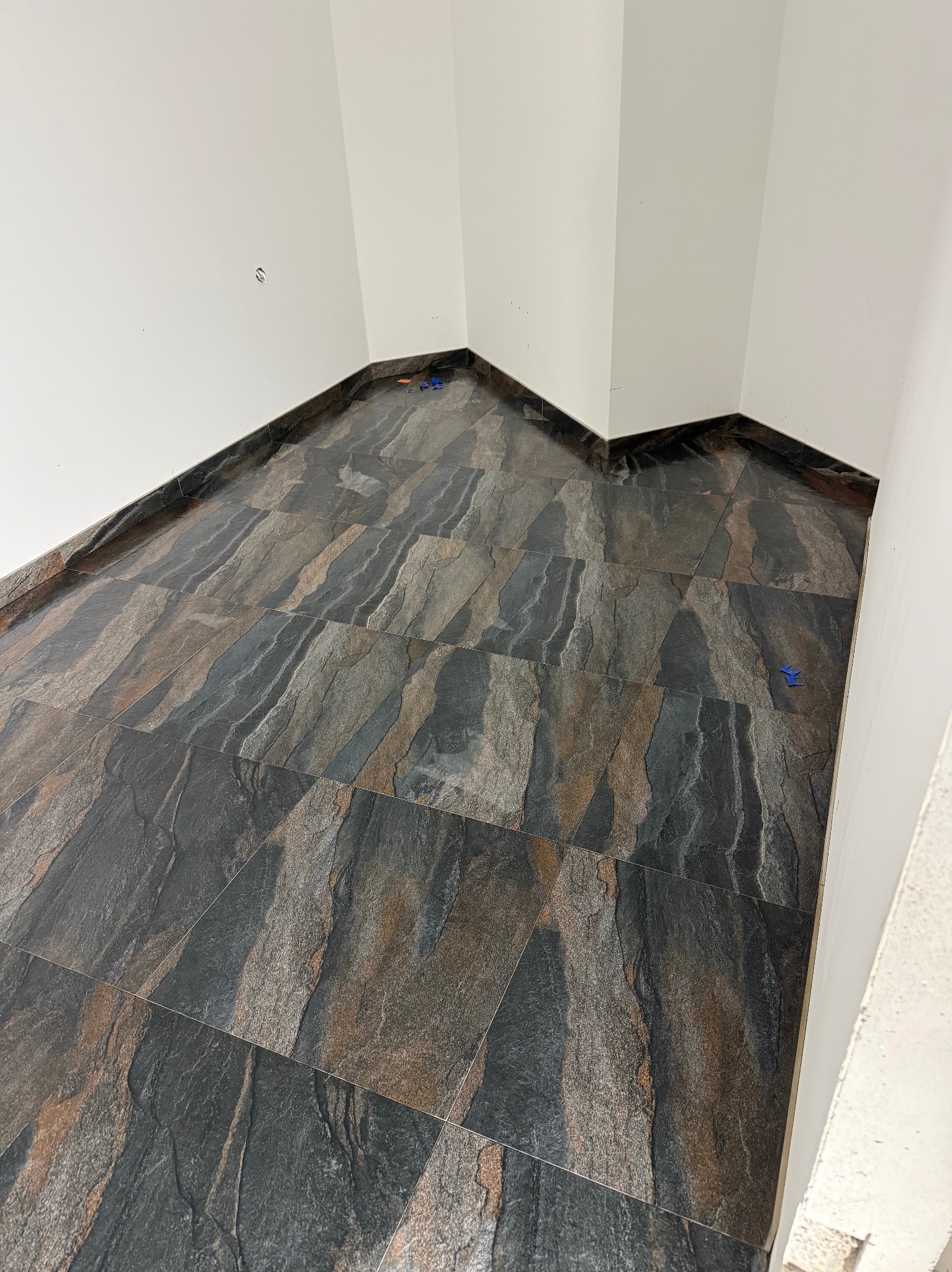 Ukończone układanie płytek podłogowych imitujących kamień w rogu pomieszczenia z białymi ścianami. Widoczne detale fug i faktura płytek.