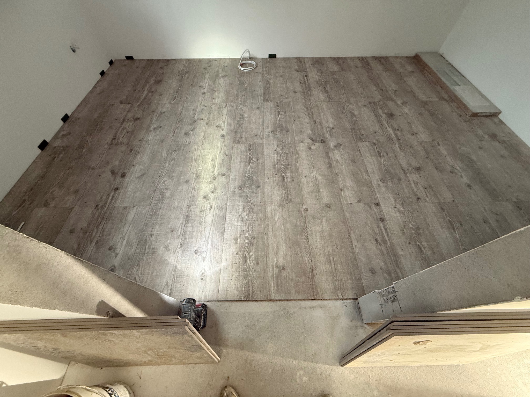 Układanie paneli podłogowych w jasnym odcieniu drewna, widoczne czarne kliny dystansowe przy ścianach, narzędzia i panele w trakcie montażu.