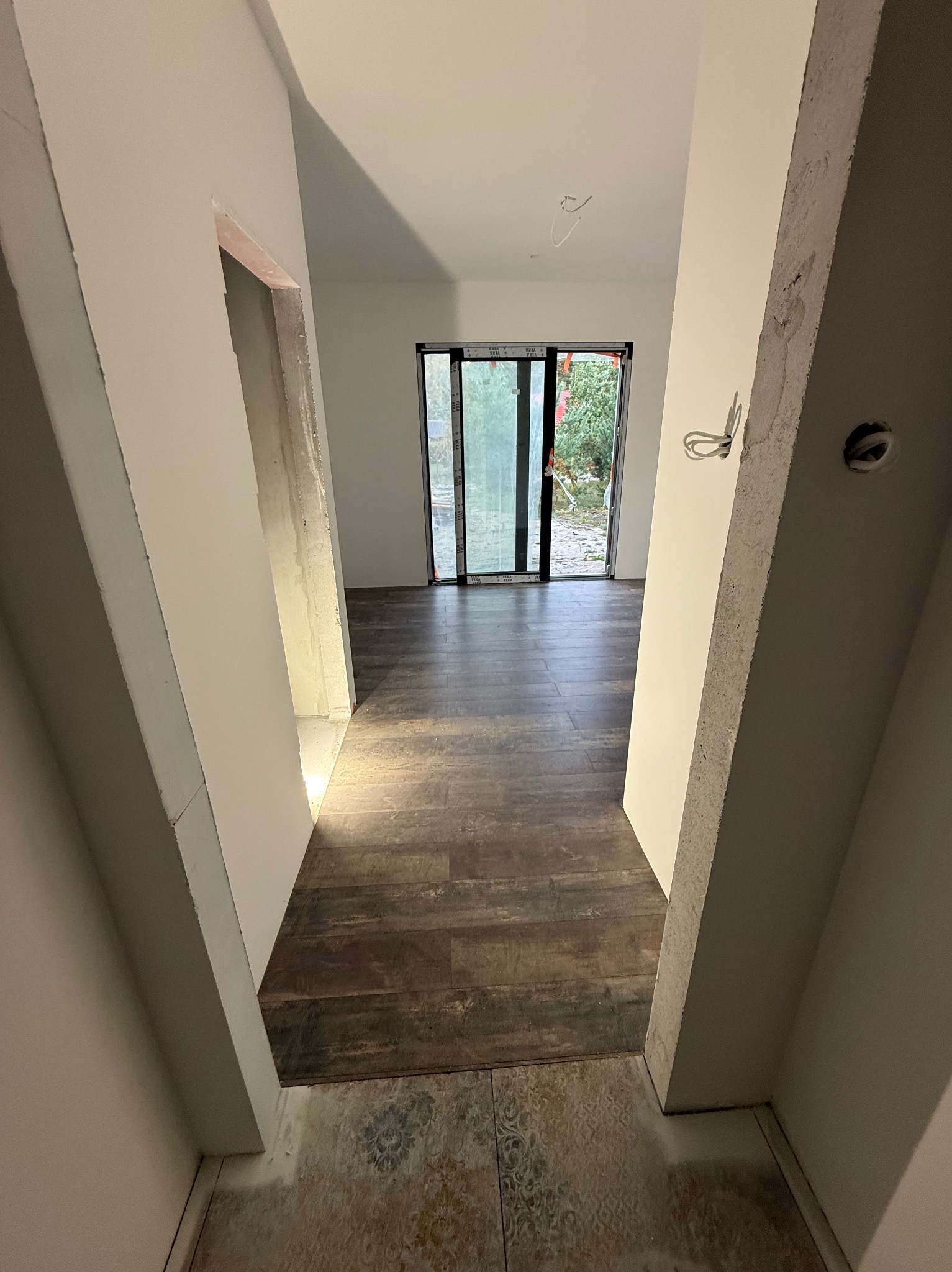 Wnętrze domu w trakcie wykańczania: ciemne panele podłogowe, surowe ściany z widocznymi przewodami elektrycznymi, okno z widokiem na zieleń. Perspektywa z korytarza.