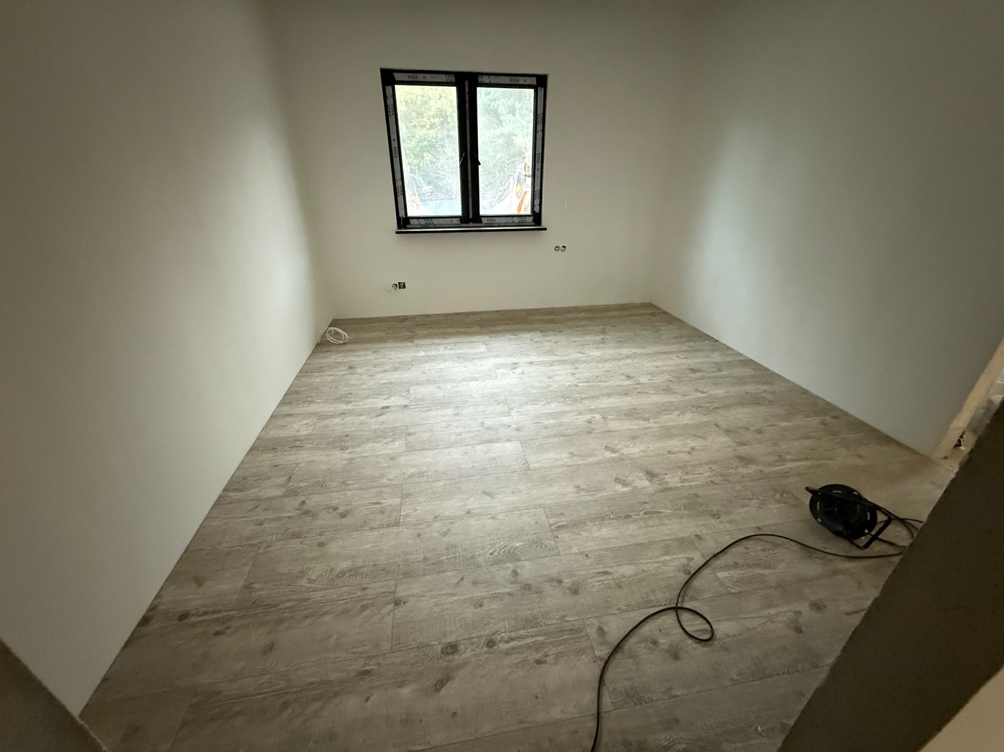 Jasne panele podłogowe ułożone w pustym pokoju z oknem. Widoczne gniazdka elektryczne na ścianie i zwinięty kabel na podłodze. Ukończony montaż paneli.