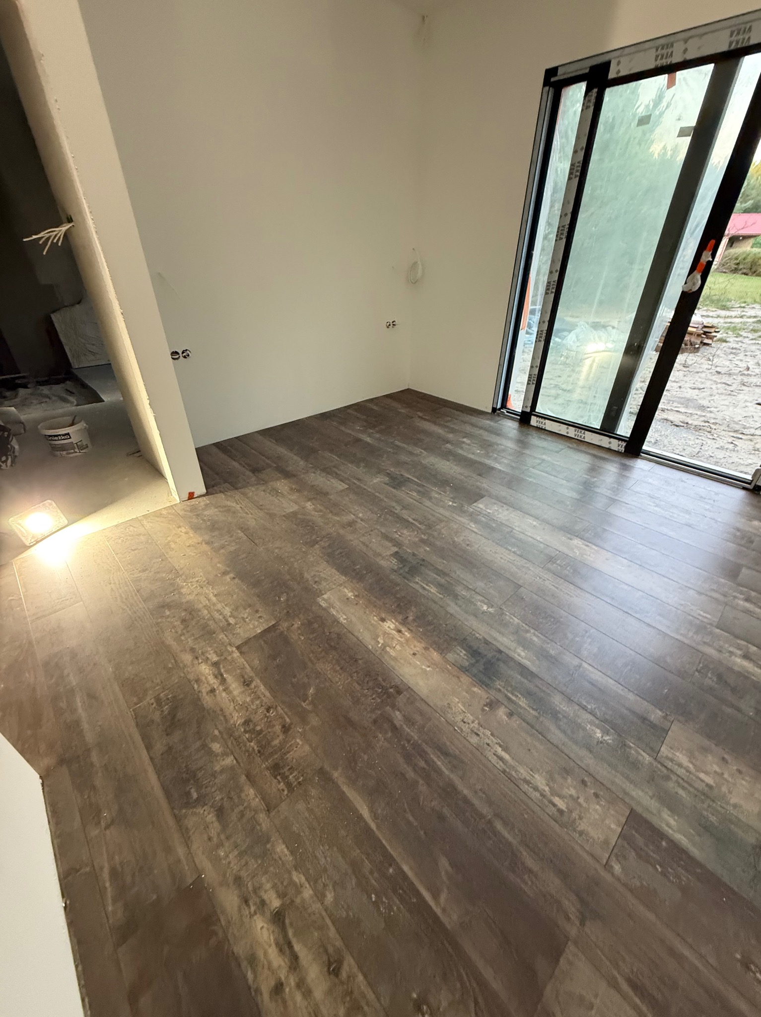 Ułożone panele podłogowe w nowym domu z dużym oknem. Widoczne detale tekstury paneli i fragment niedokończonej ściany. Naturalne światło wpadające przez okno.