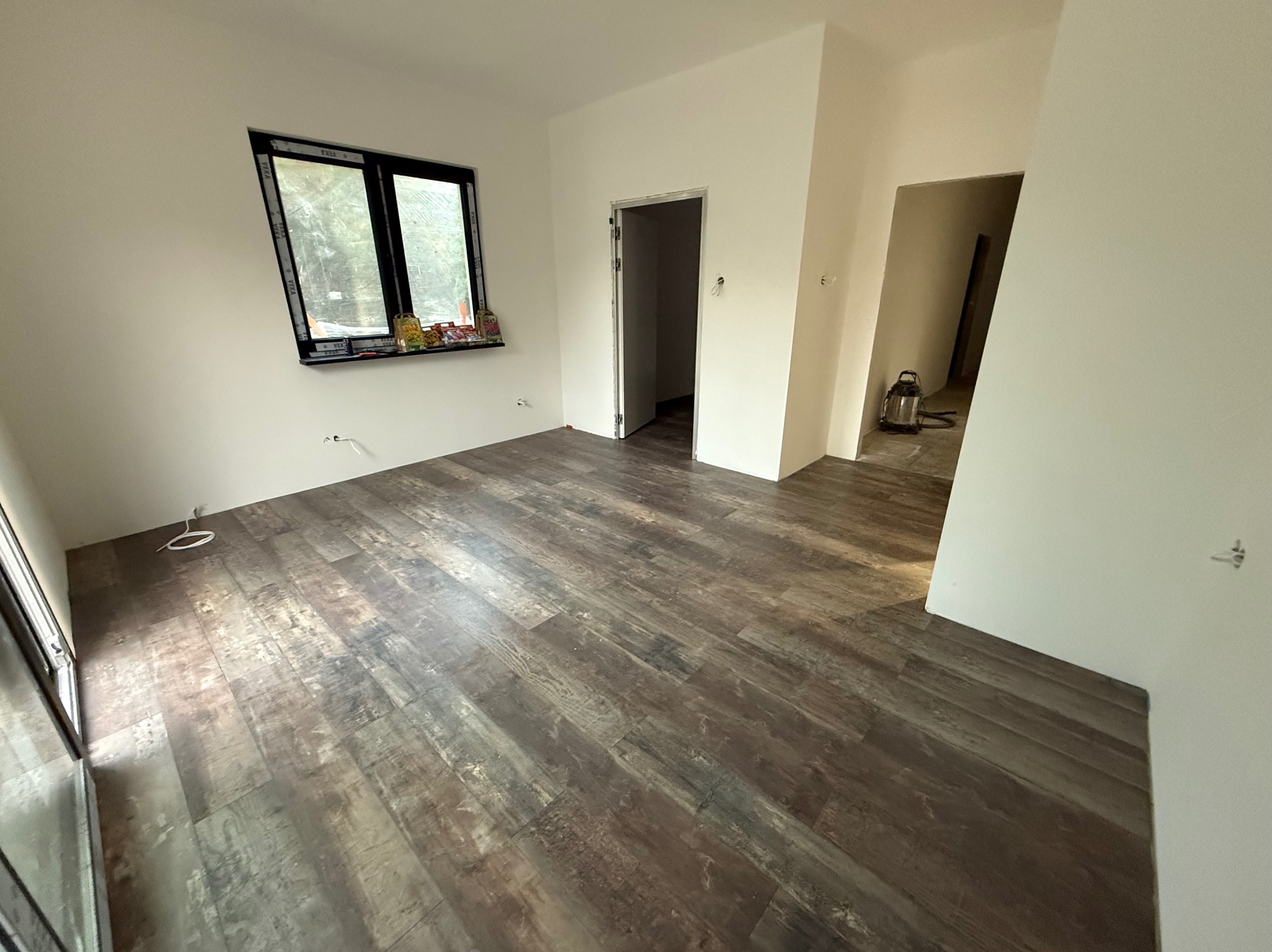 Jasne wnętrze po remoncie z panelami podłogowymi w odcieniach brązu. Widoczne białe ściany, okno i otwory drzwiowe. W tle odkurzacz.