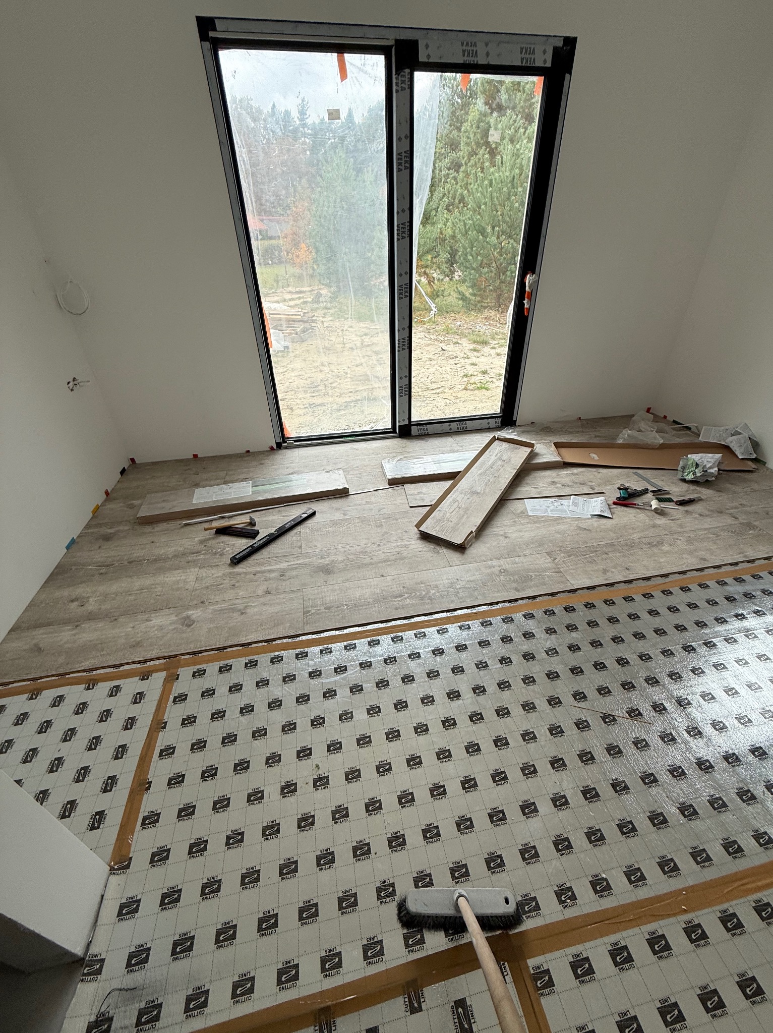 Układanie paneli podłogowych w pokoju z oknem dachowym. Widoczna folia izolacyjna, panele i narzędzia. Częściowo ułożona podłoga z paneli imitujących drewno.