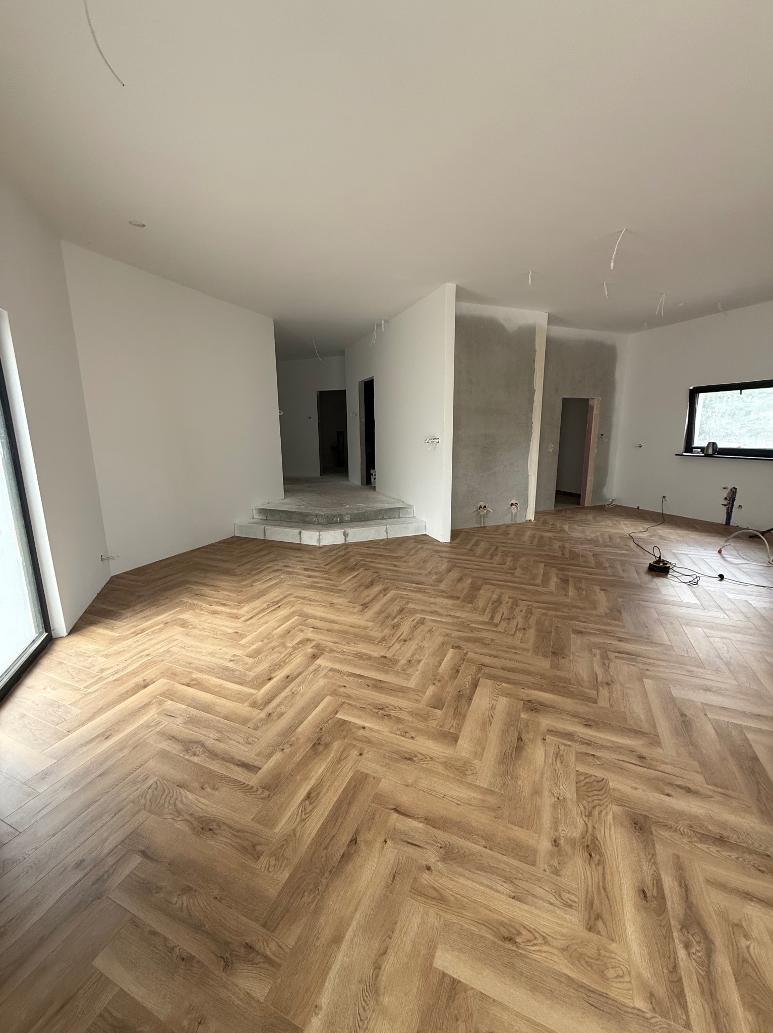 Jasne panele ułożone we wzór jodełki francuskiej w przestronnym, częściowo wykończonym wnętrzu. Widoczne białe ściany, surowy beton i wystające kable.