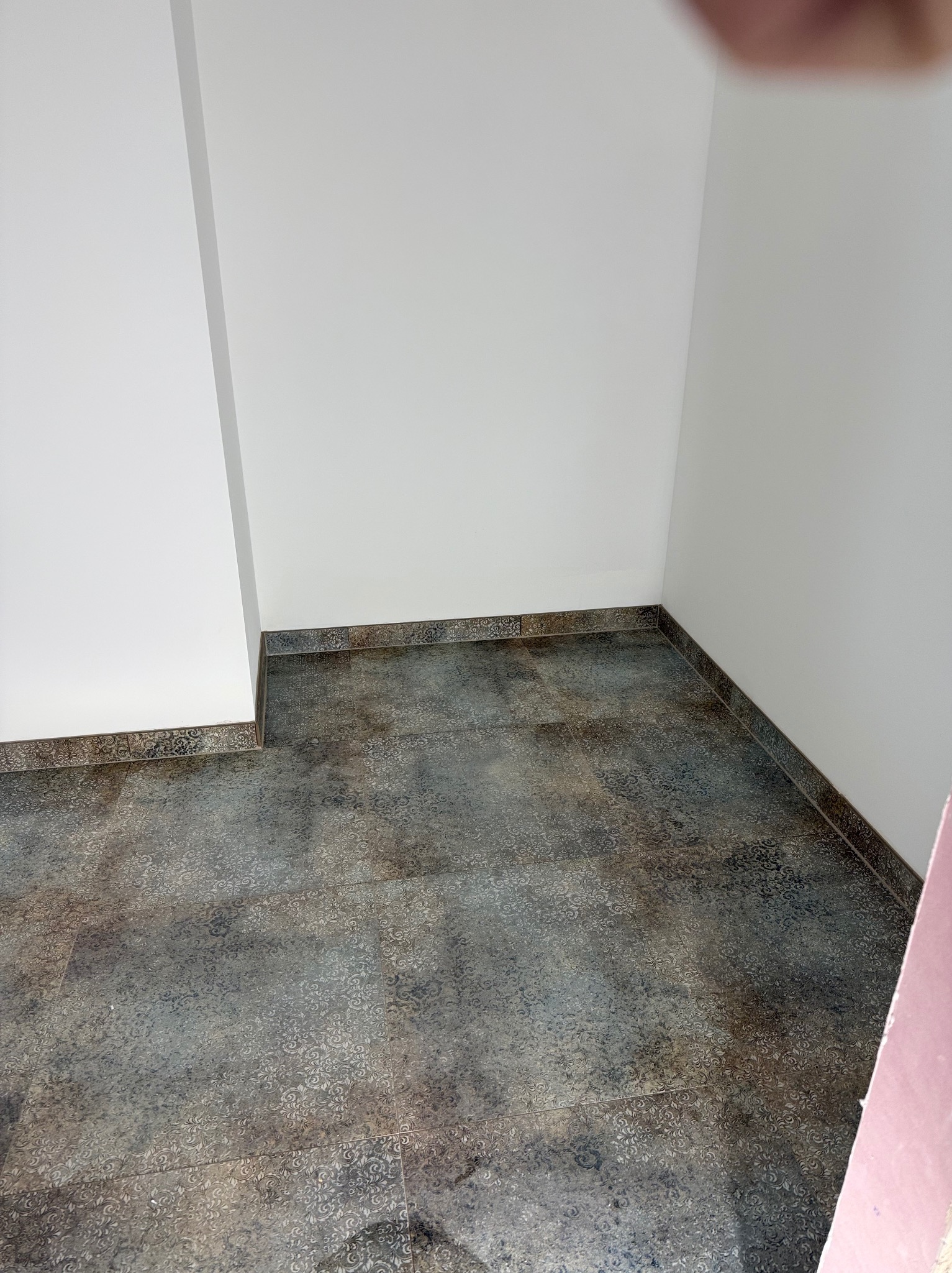 Wykończone wnętrze z ciemną, wzorzystą podłogą z płytek i białymi ścianami. Widoczne listwy przypodłogowe z dekoracyjnym motywem. Ujęcie z narożnika pomieszczenia.