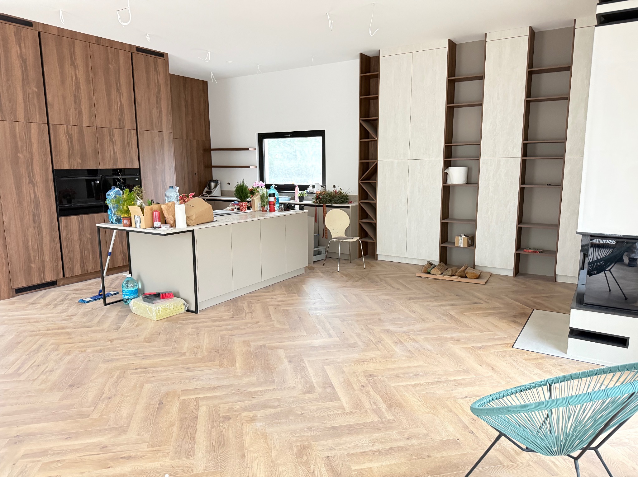 Jasna, przestronna kuchnia z parkietem ułożonym w jodełkę. Widoczne zabudowy meblowe, wyspa kuchenna i elementy dekoracyjne. Całość w nowoczesnym stylu.