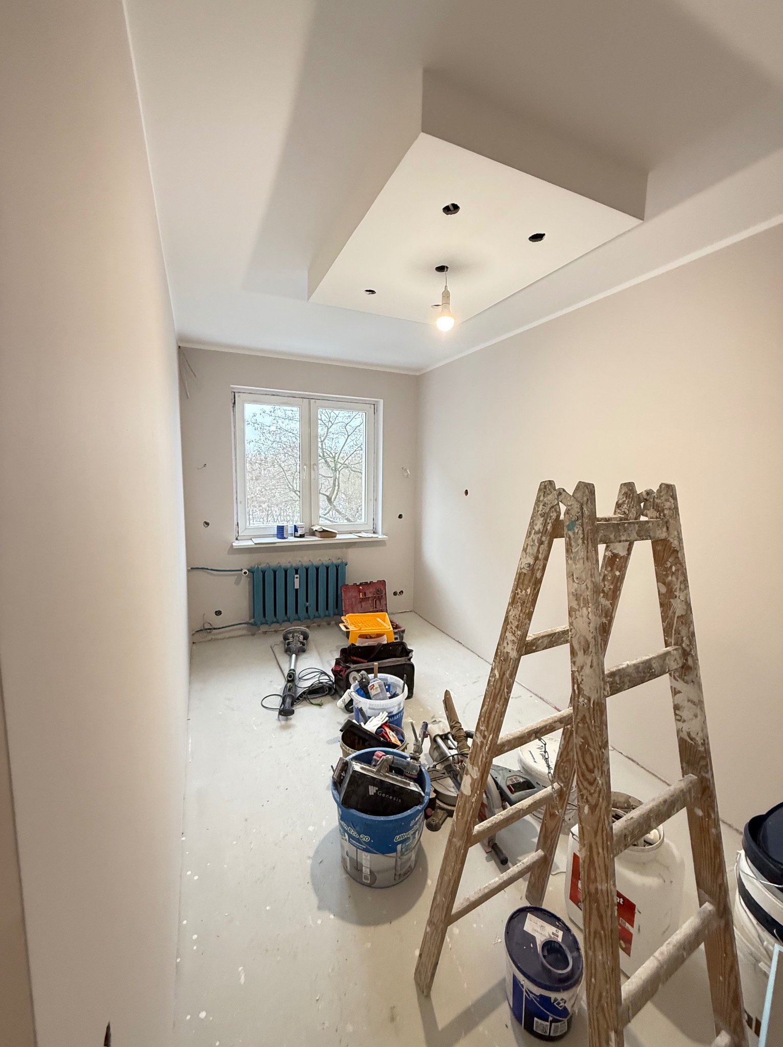 Pokój po szpachlowaniu, przygotowany do malowania. Widoczne narzędzia, drabina malarska i resztki gładzi na podłodze. Ściany gładkie, w jasnym odcieniu.