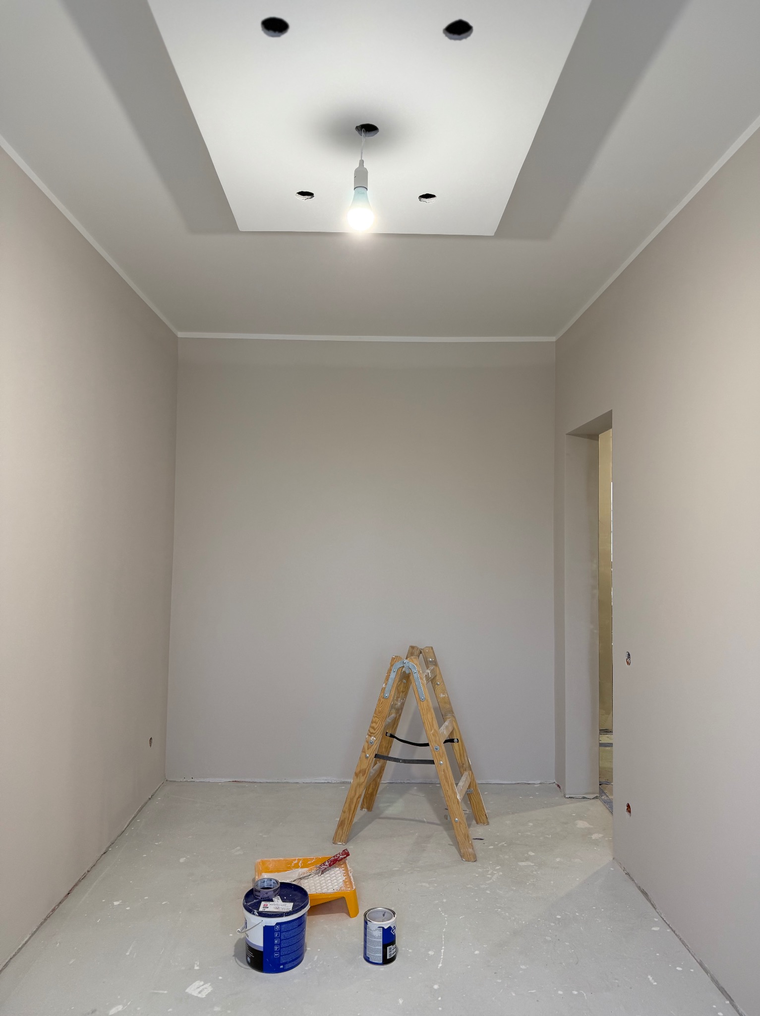 Wnętrze po malowaniu ścian na neutralny odcień szarości. Widoczna drabina, puszki z farbą i kuweta malarska. Sufit z podwieszanym elementem i oświetleniem.