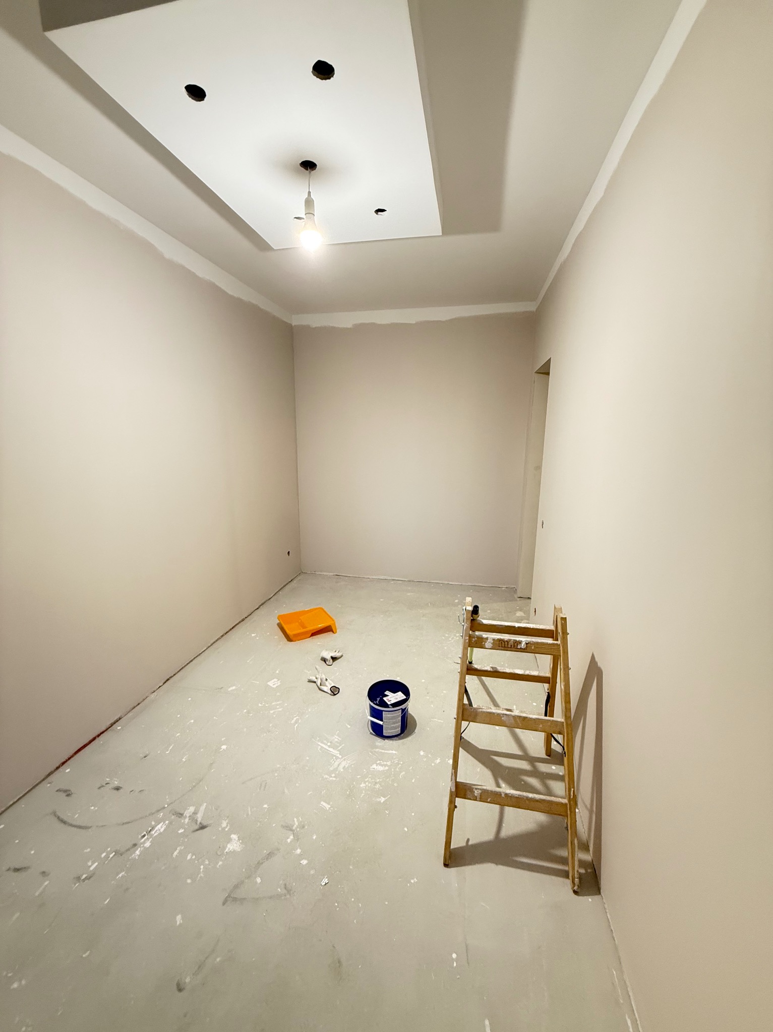 Jasne wnętrze w trakcie odświeżania: ściany pomalowane na beżowo, widoczna drabina, kuweta malarska i puszka z farbą. Sufit z oświetleniem w centralnej części.