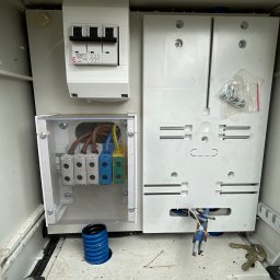 3Miejski Elektryk - Otwarta, brudna skrzynka elektryczna z widocznymi bezpiecznikami, złączkami kablowymi pod przezroczystą osłoną, niebieską rurą karbowaną u dołu i kluczami leżącymi na dnie.