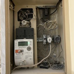 3Miejski Elektryk - Wnęka z zamontowanym licznikiem energii elektrycznej Sagemcom CX1000-6 S, zabezpieczeniami na szynie DIN, gniazdem elektrycznym i przewodami wewnątrz obudowy z widocznymi śladami użytkowania.