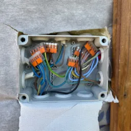 Otwarta puszka elektryczna z widocznymi przewodami połączonymi pomarańczowymi złączkami WAGO, zamontowana na ścianie z izolacją i drewnianą deską.