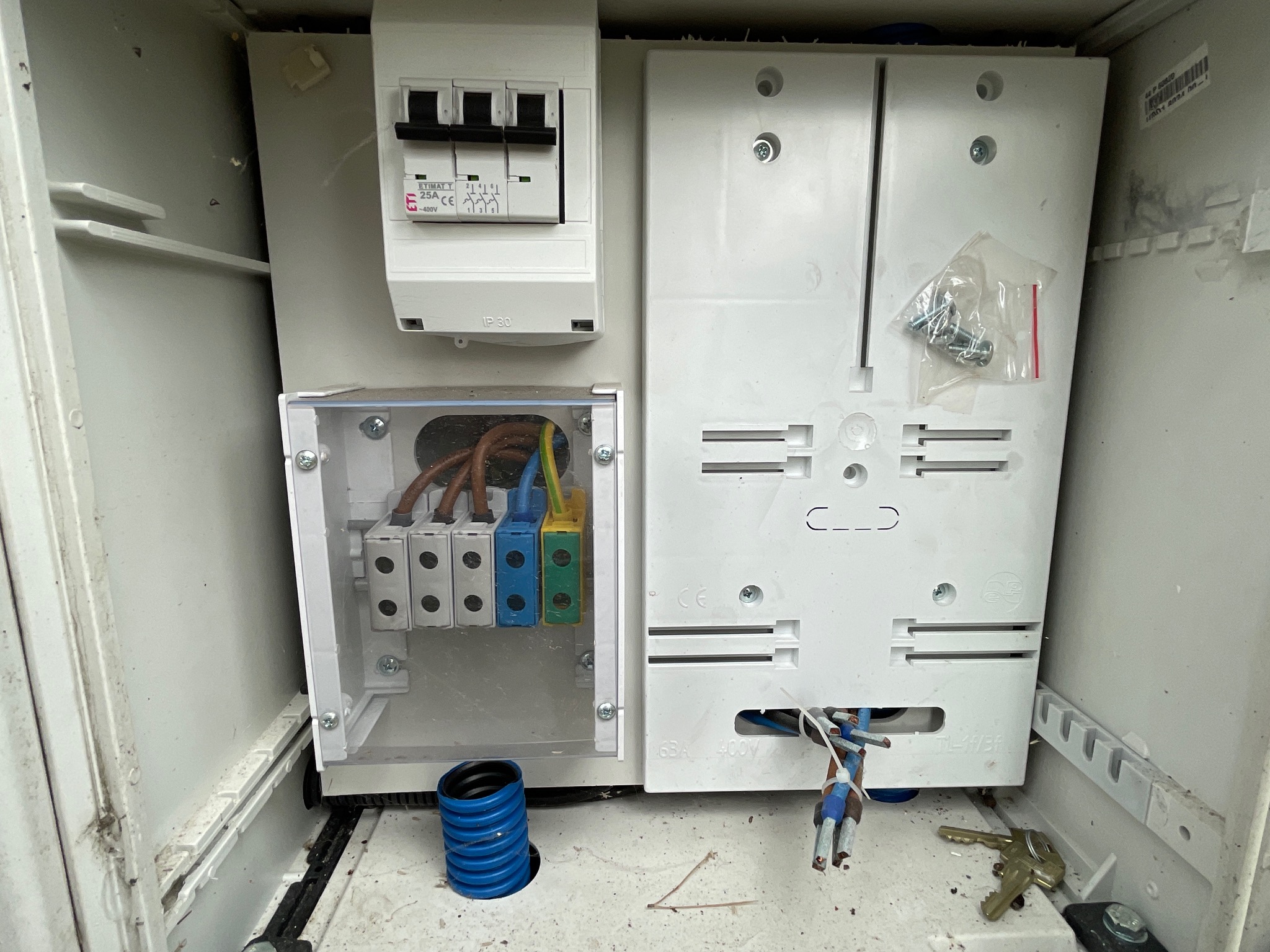 Otwarta, brudna skrzynka elektryczna z widocznymi bezpiecznikami, złączkami kablowymi pod przezroczystą osłoną, niebieską rurą karbowaną u dołu i kluczami leżącymi na dnie.