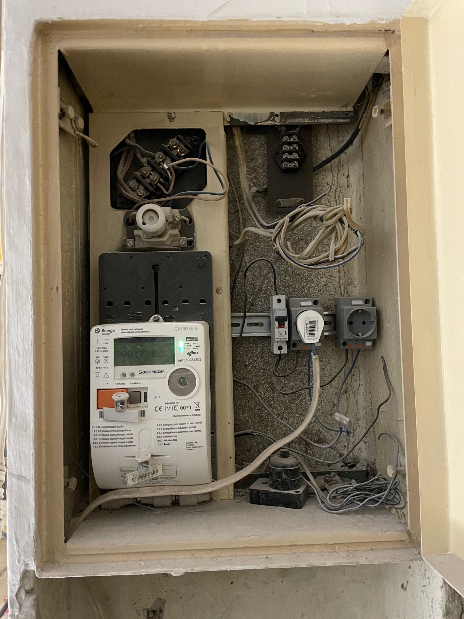 Wnęka z zamontowanym licznikiem energii elektrycznej Sagemcom CX1000-6 S, zabezpieczeniami na szynie DIN, gniazdem elektrycznym i przewodami wewnątrz obudowy z widocznymi śladami użytkowania.