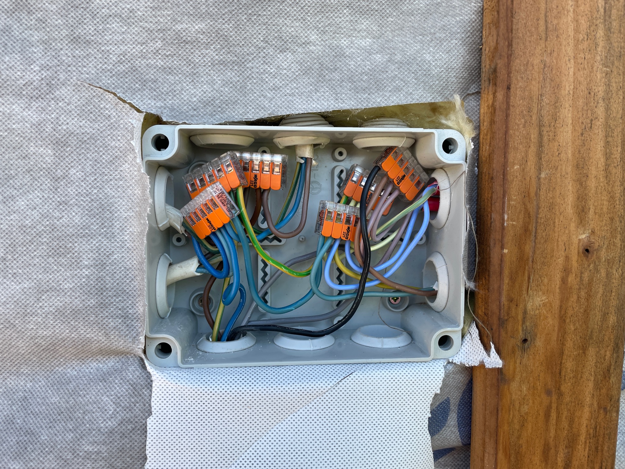 Otwarta puszka elektryczna z widocznymi przewodami połączonymi pomarańczowymi złączkami WAGO, zamontowana na ścianie z izolacją i drewnianą deską.
