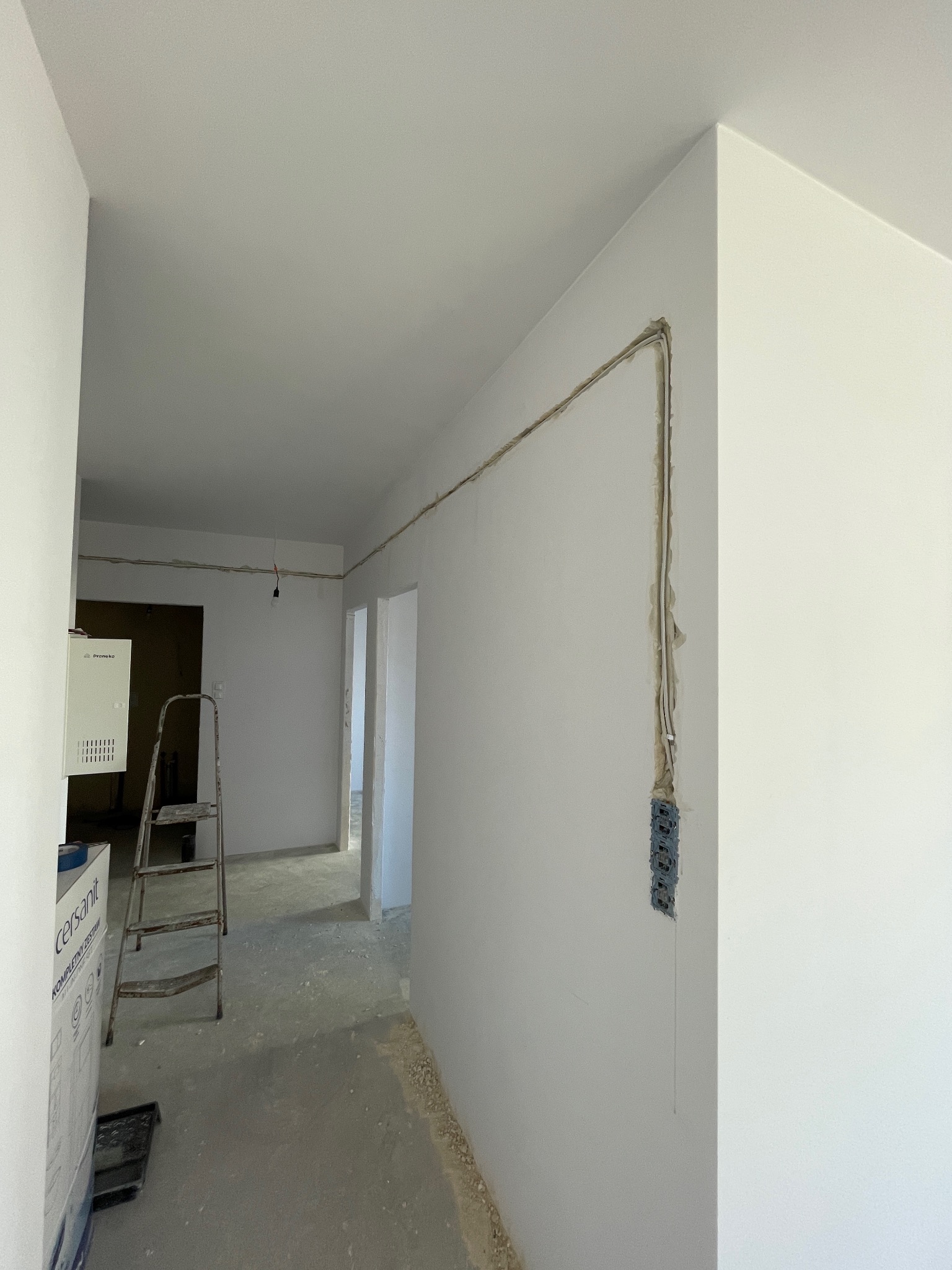 Wnętrze w trakcie remontu z widocznymi przewodami elektrycznymi poprowadzonymi po ścianach, puszki pod gniazdka, drabina i karton z materiałami budowlanymi w tle.