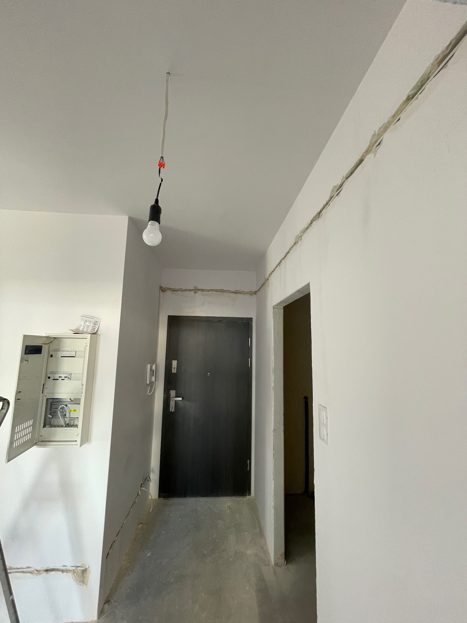 Wnętrze w trakcie remontu: wisząca żarówka na kablu, otwarta skrzynka elektryczna na ścianie, ślady po przewodach na suficie i ścianach, przygotowane do malowania.
